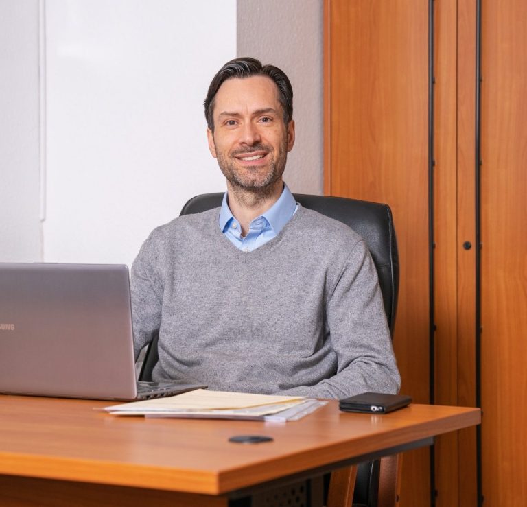 Wir sind für sie da Ein Mann sitzt an einem Schreibtisch, lächelt und arbeitet am Laptop.